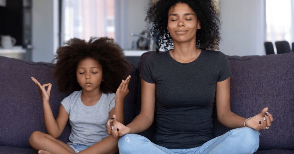 A mom and adolescent daughter sitting on couch cross-legged with eyes closed practicing meditation to reduce stress and chronic inflammation.