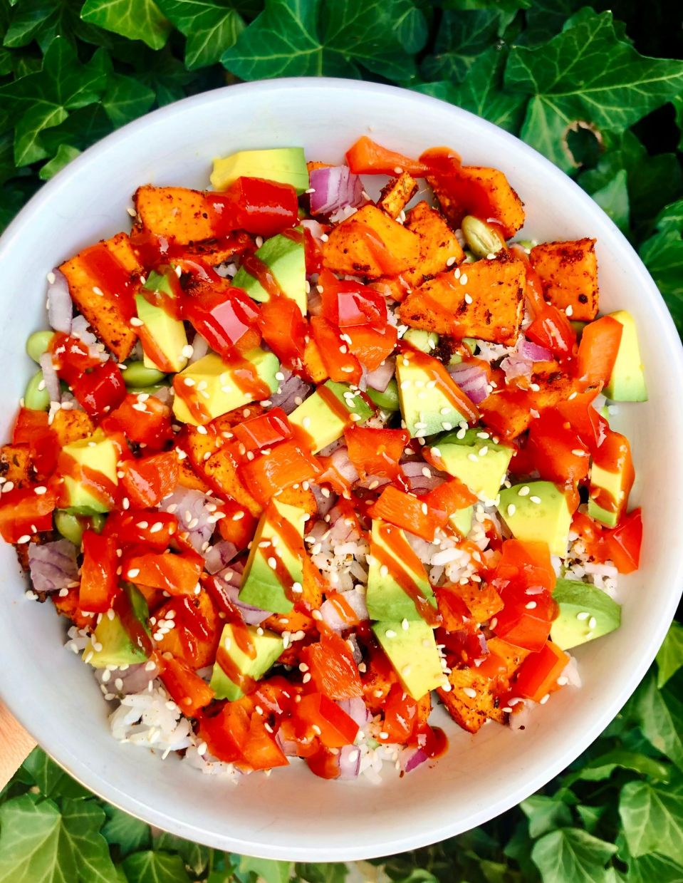 Healthy rice bowl with edamame, sweet potato, avocado, and colorful vegetables, topped with sesame seeds and a drizzle of sriracha sauce.