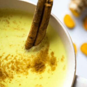 A warm cup of golden milk made with turmeric and sprinkled with cinnamon, served in a ceramic mug with a side of fresh turmeric and cinnamon sticks.