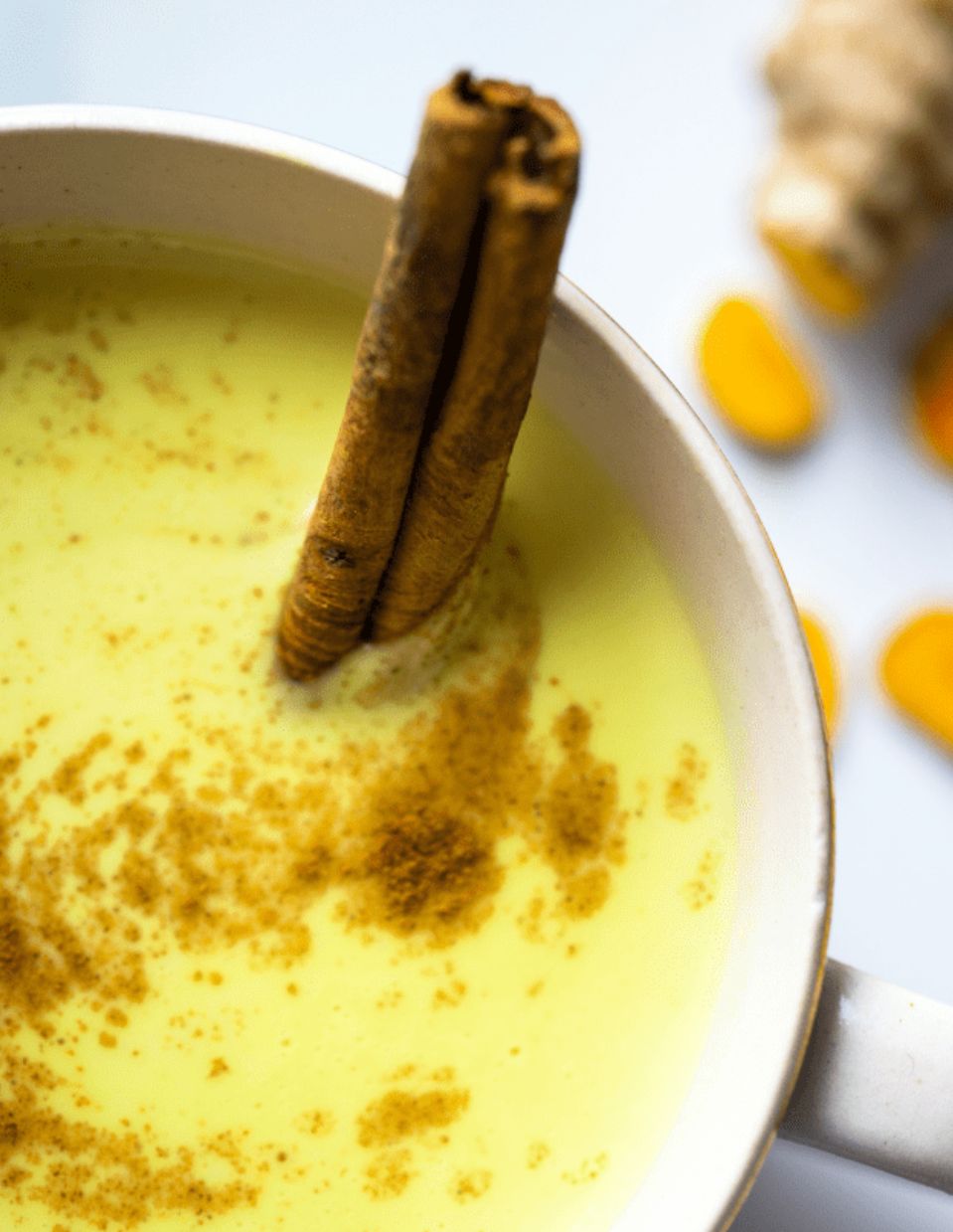 A warm cup of golden milk made with turmeric and sprinkled with cinnamon, served in a ceramic mug with a side of fresh turmeric and cinnamon sticks.