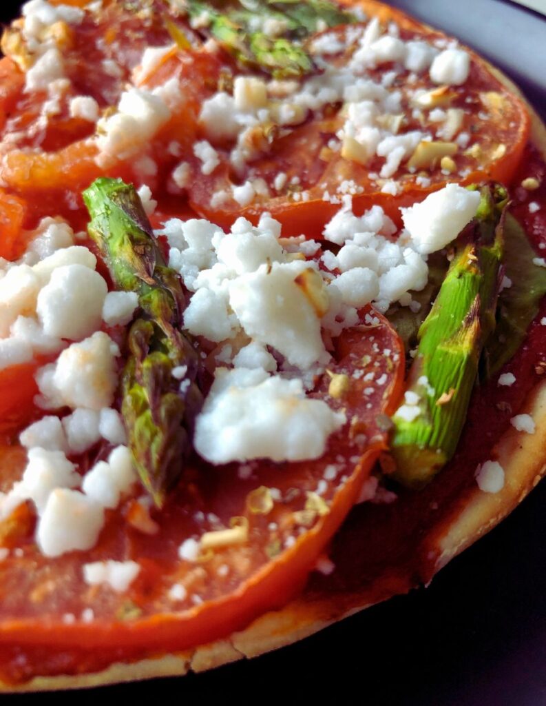 Veggie pizza topped with asparagus, tomatoes, garlic, and feta on a crispy pita crust, served fresh from the oven.