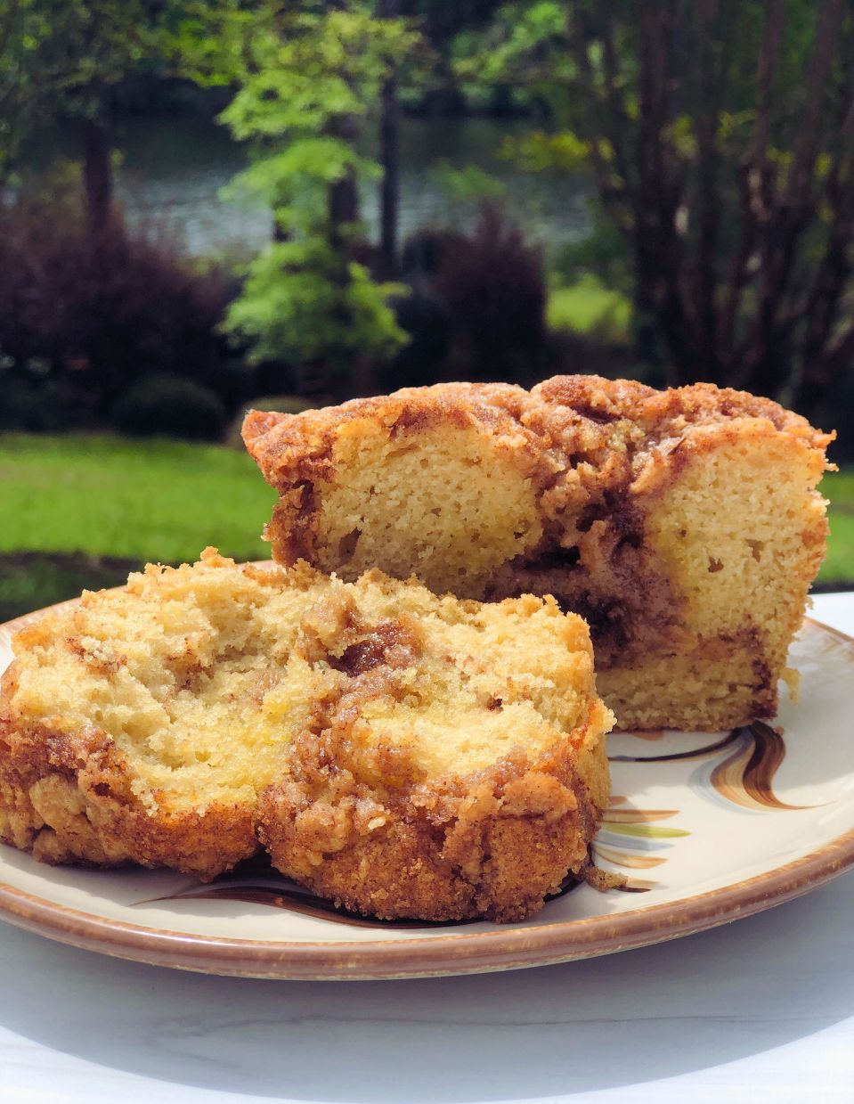 A slice of vegan Cinnamon Streusel Coffee Cake on a beautifully hand-painted Gail Pittman plate, set outdoors on a sunny day, surrounded by lush greenery.