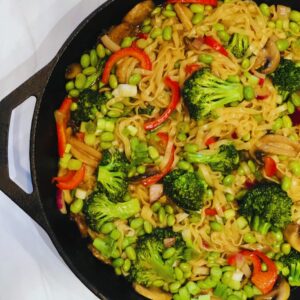 Vegan asian stir fry recipe made in a cast iron skillet with noodles, colorful broccoli mushrooms, and red bell peppers mixed with a savory sauce garnished with green onions.