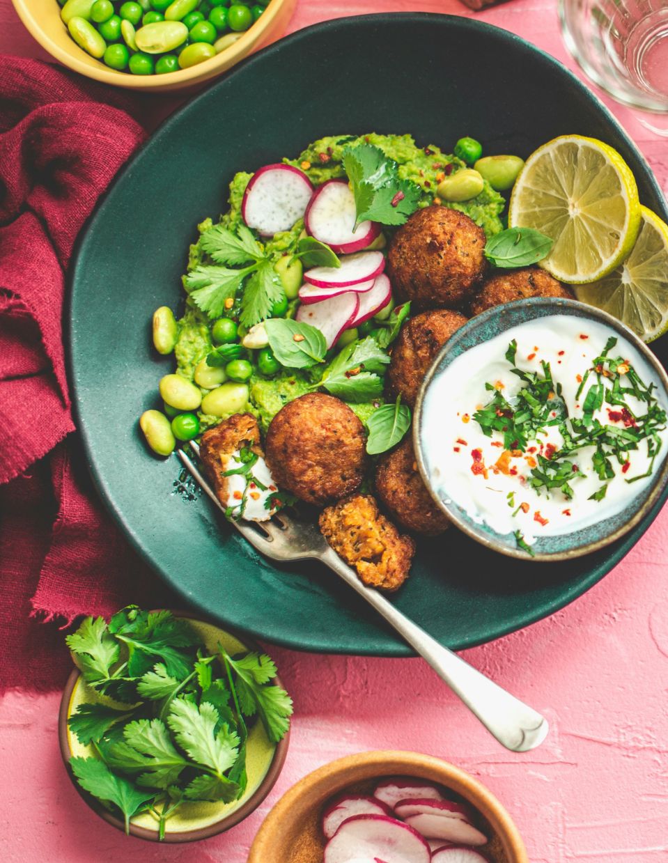 A vibrant plant-based, weight loss meal featuring falafel, green pea mash, fresh radish slices, edamame, and herbs, served with a side of creamy yogurt sauce and garnished with lemon wedges.