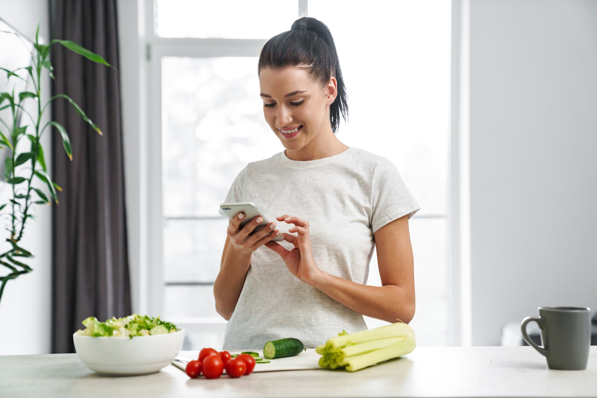 Woman in her kitchen with healthy foods on the counter, looking at her phone while receiving telehealth nutrition counseling from a registered dietitian at home.