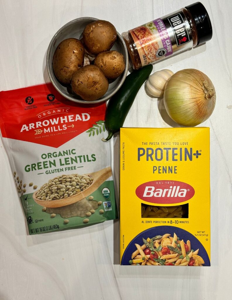 Ingredients for a high-protein, healthy vegan Cajun pasta recipe, including Barilla Protein Plus Penne Pasta, Weber New Orleans Cajun Seasoning, Portobello mushrooms, sweet onion, garlic cloves, jalapeño, and Arrowhead Mills green lentils.