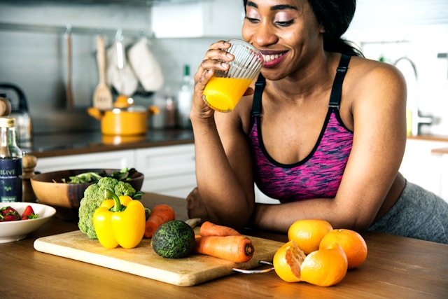 A woman standing in a modern kitchen drinking a turmeric ginger shot with fruits and vegetables on the counter, representing holistic nutrition for weight loss and health.