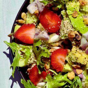 A vibrant Strawberry Walnut Salad with fresh strawberries, avocado, walnuts, red onion, and organic spring mix, drizzled with balsamic dressing and served in on a black plate.