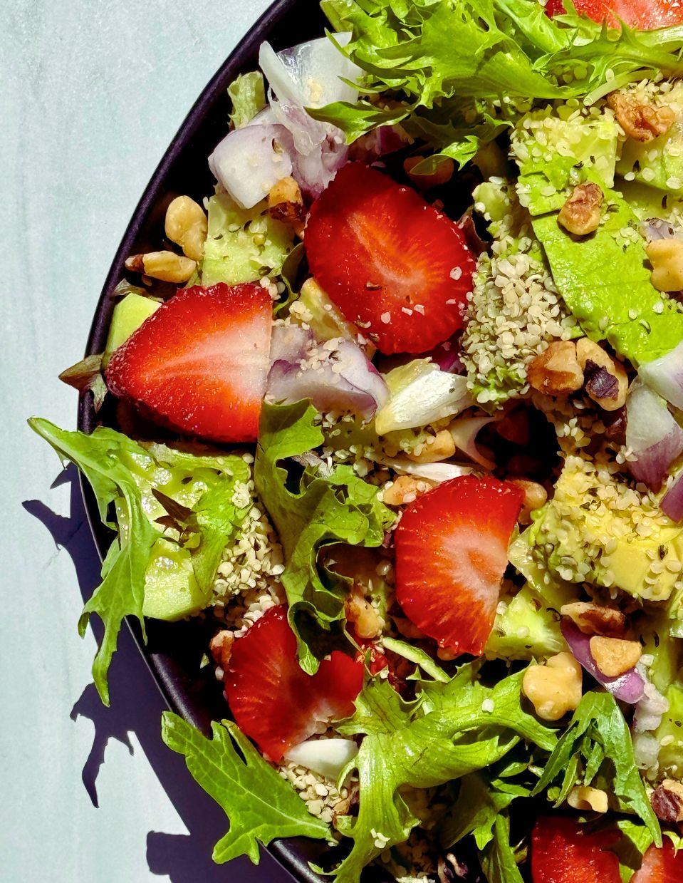 A vibrant Strawberry Walnut Salad with fresh strawberries, avocado, walnuts, red onion, and organic spring mix, drizzled with balsamic dressing and served in on a black plate.