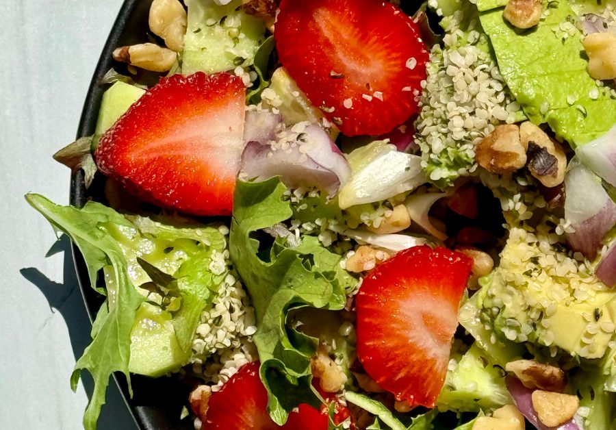 Close-up view of fresh strawberries, walnuts, avocado slices, red onion, and hemp seeds on a black plate.