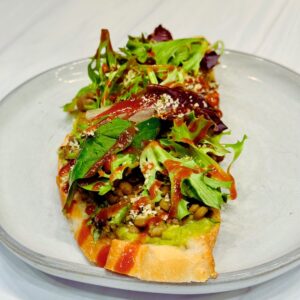 Avocado toast topped with guacamole, cooked green lentils, spring mix lettuce, hemp hearts, and a drizzle of sriracha sauce on a slice of toasted sourdough bread for a easy, high protein, healthy vegan meal.