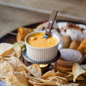 A creamy vegan nacho cheese dip in a bowl, placed on a tray alongside tortilla chips and a variety of healthy appetizers.